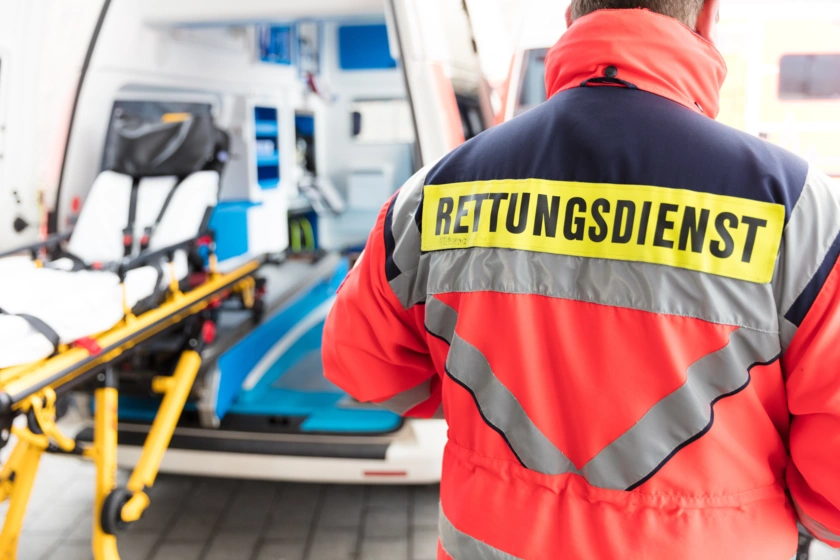 Rettungssanitäter steht mit "Rettungsdienst" beschrifteter Jacke auf dem Rücken neben einem Krankenwagen. Eine Krankentrage ist am geöffneten Krankenwagen ersichtlich.